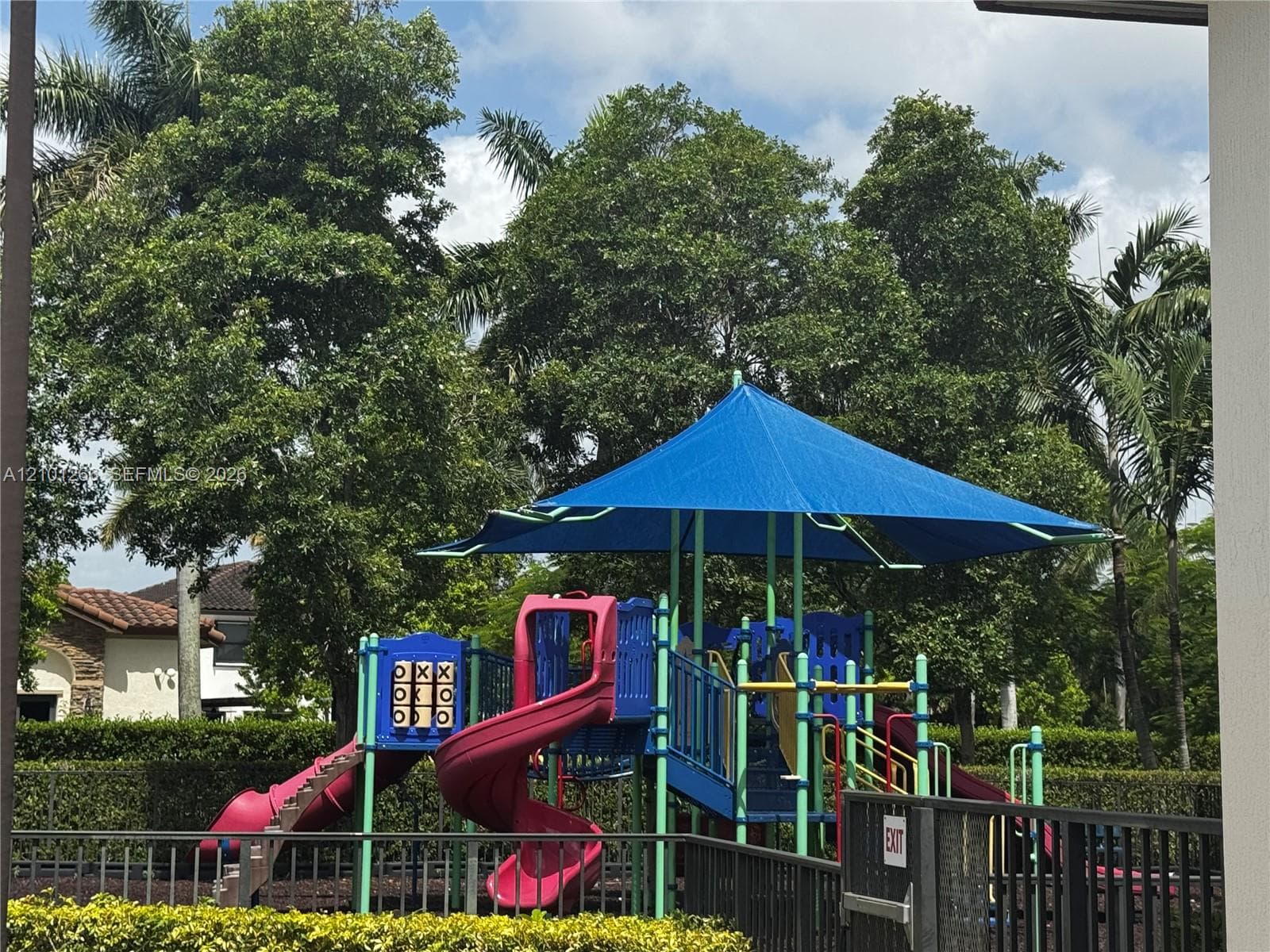 Outdoor recreational structure with a blue shade canopy, integrated spiral slide, and game panel, enclosed by perimeter fencing and surrounded by mature trees
