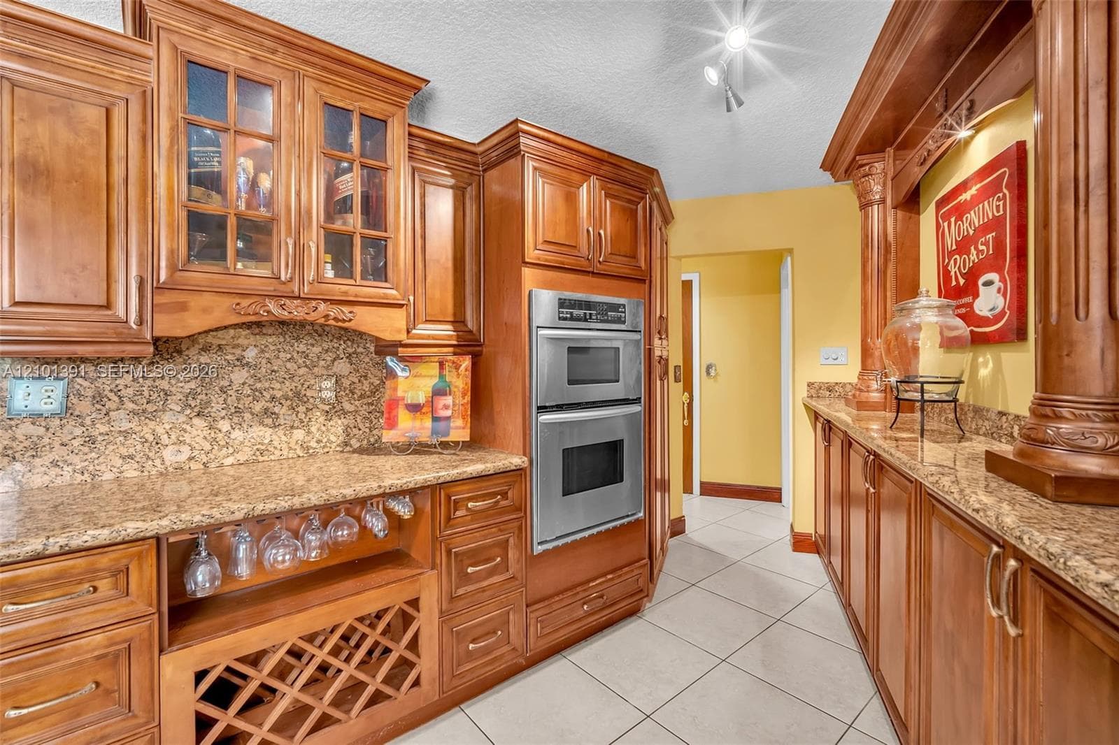 Custom wood cabinetry with glass-front accents, stainless steel double wall ovens, a built-in wine rack, and a stone-finish backsplash with countertops