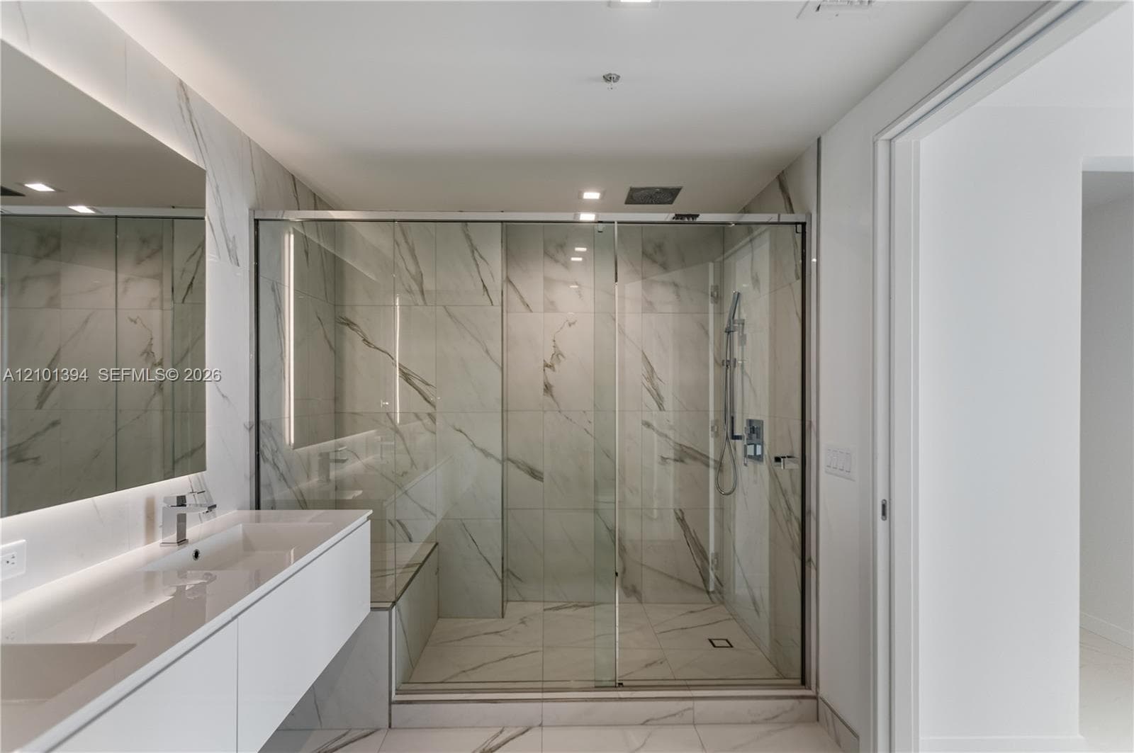 Floating white vanity with integrated dual sinks, large frameless wall-mounted mirror, and walk-in glass-enclosed shower featuring stone-finish wall and floor tile