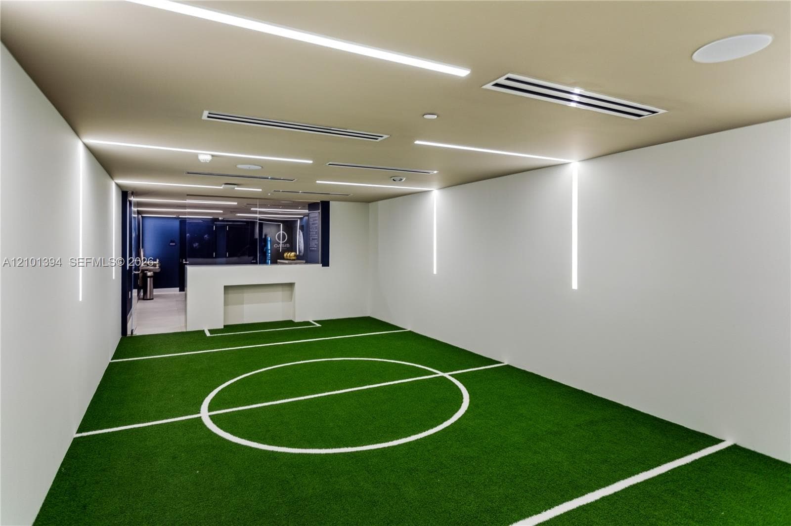 Indoor recreation area featuring synthetic turf flooring with integrated field markings, recessed linear ceiling lighting, vertical wall-mounted accent lighting, and a built-in counter