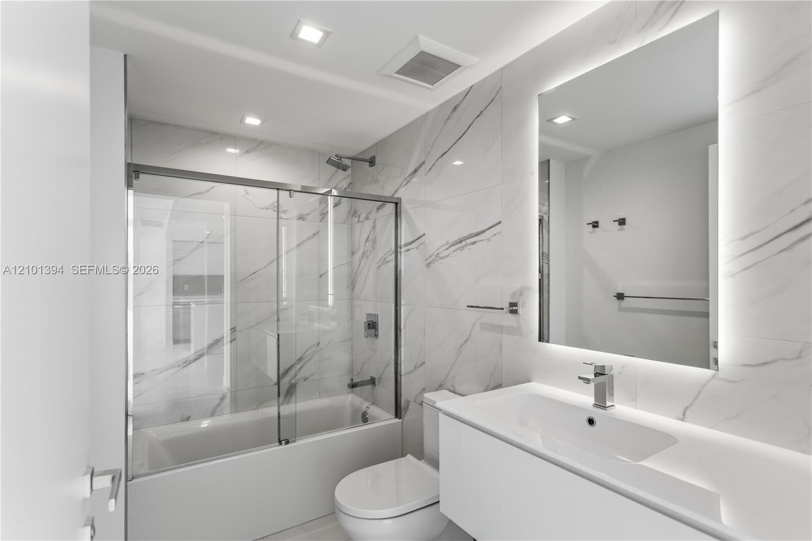 Full-height veined stone-look wall tiling, white floating vanity with integrated sink, LED-backlit vanity mirror, and glass-enclosed shower and tub combination with chrome fixtures
