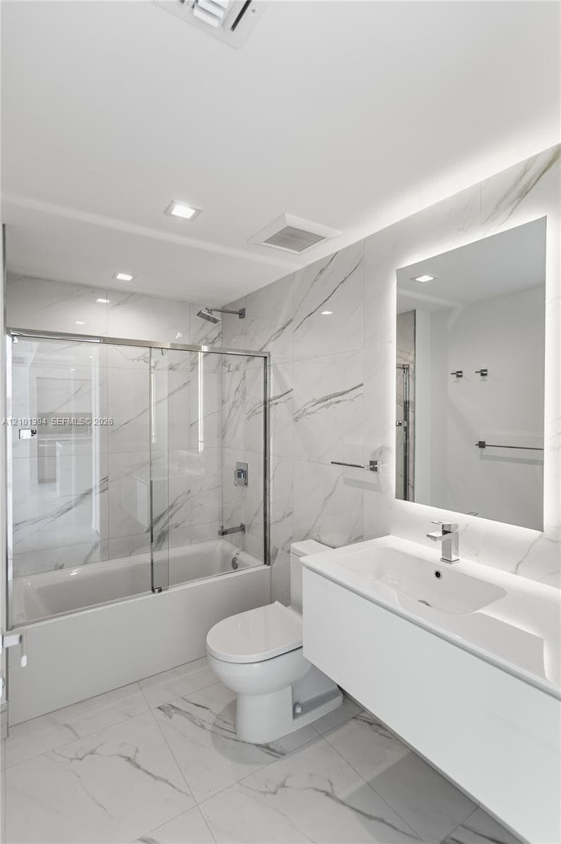Floating vanity with integrated sink, backlit wall mirror, glass-enclosed shower and tub combination, and stone-finish floor and wall tile
