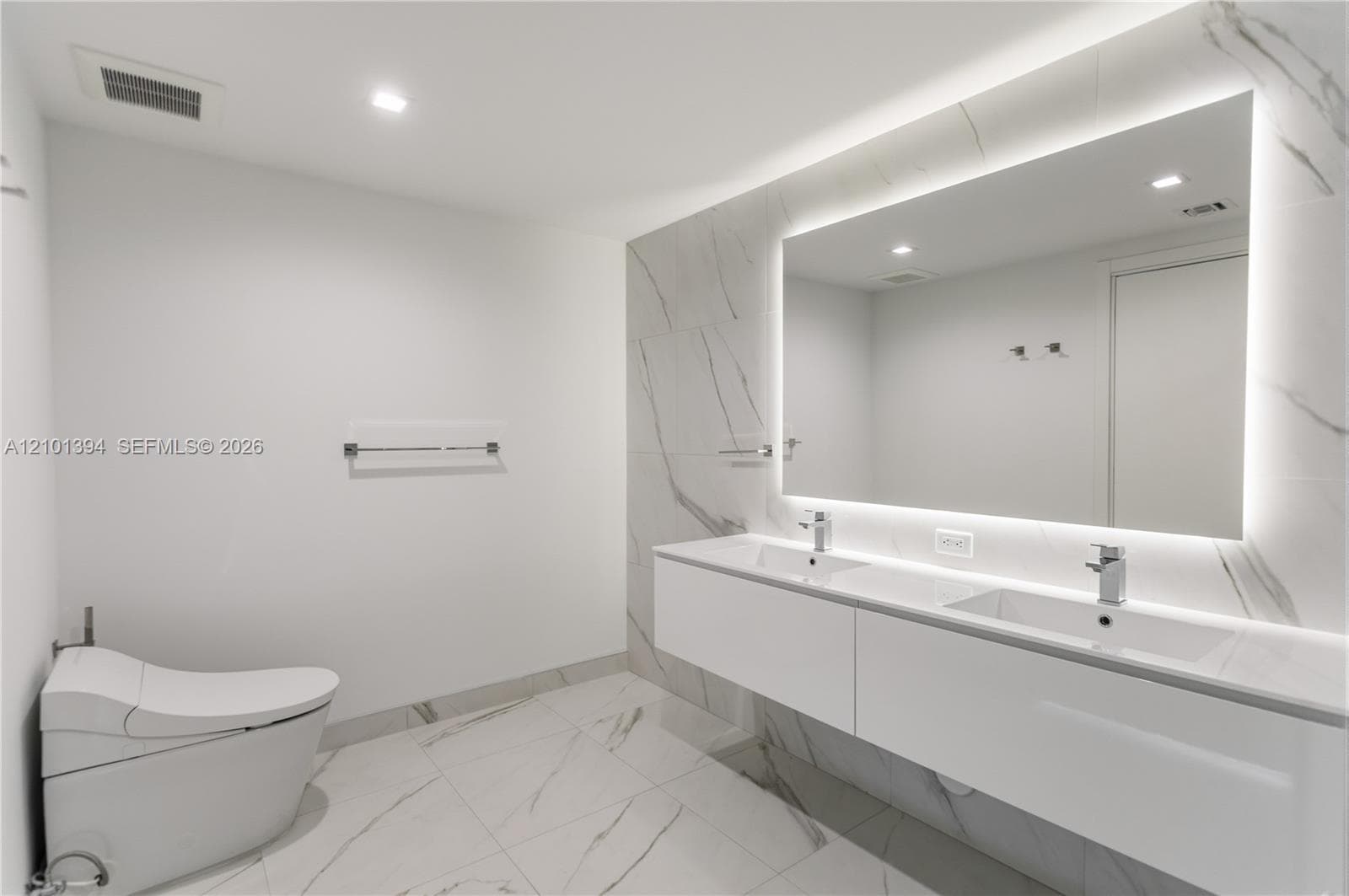 Modern floating dual-sink vanity with a solid surface countertop, large backlit mirror, and floor-to-ceiling stone-finish tile wall featuring a wall-mounted toilet
