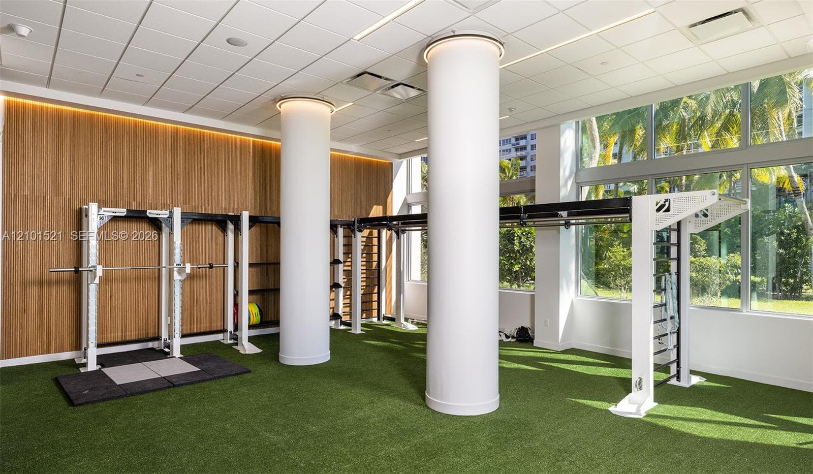 Fitness space featuring floor-to-ceiling windows, wood-paneled walls, synthetic turf flooring, recessed ceiling lighting, and structural cylindrical columns