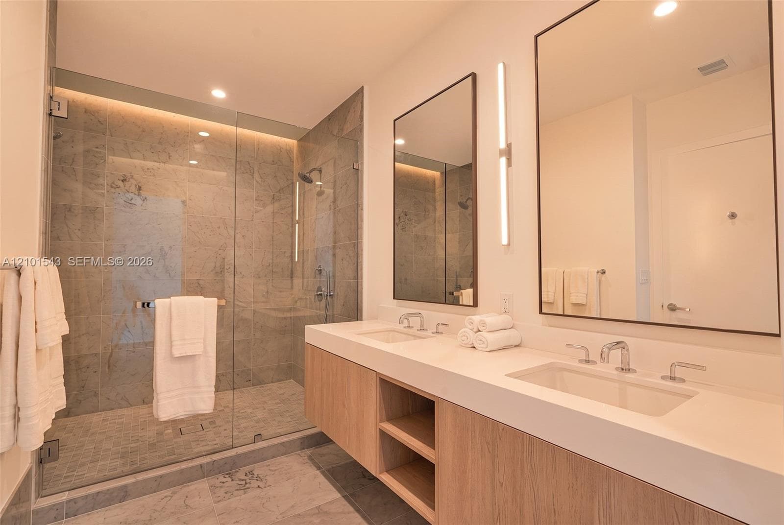 Floating dual-sink vanity with a solid surface countertop, glass-enclosed walk-in shower with stone tile surround, stone tile flooring, and integrated vertical lighting fixtures