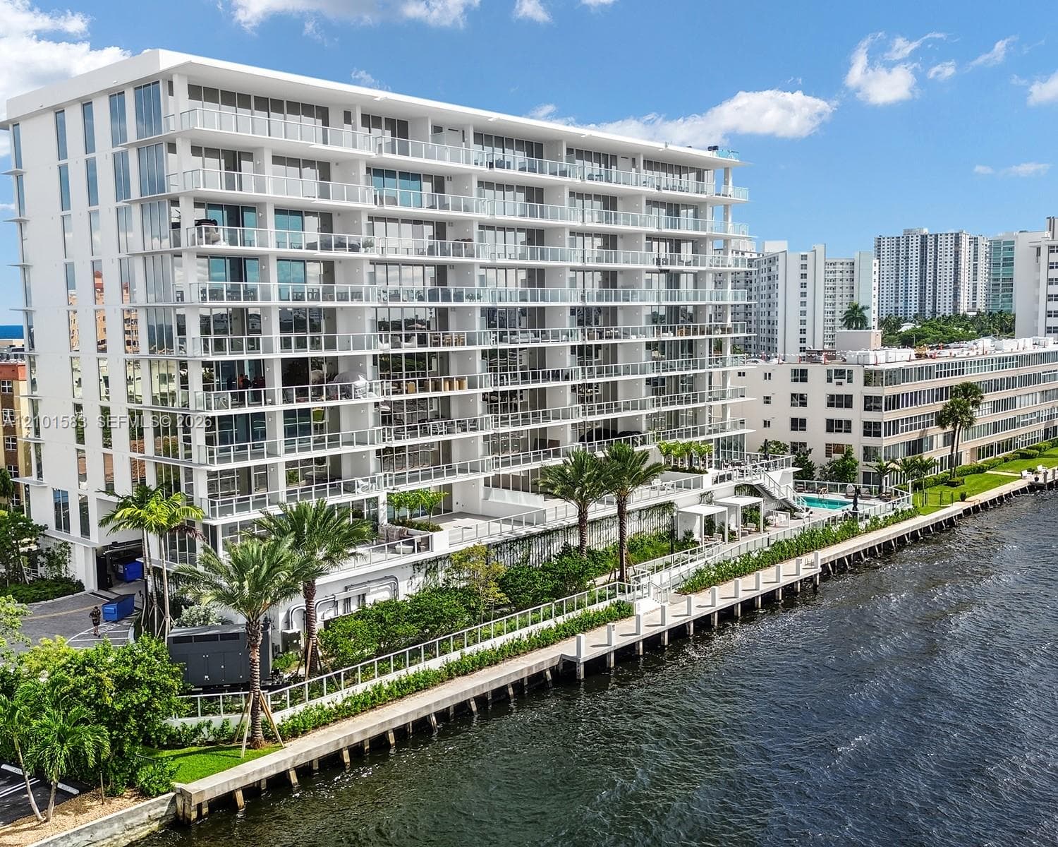 Modern mid-rise building featuring floor-to-ceiling glass windows, wraparound glass-panel balconies, a waterfront bulkhead, and a paved canal-side walkway