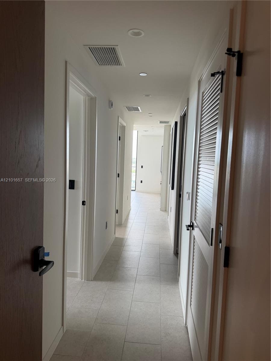 Hallway featuring large-format ceramic tile flooring, recessed ceiling lighting, and matte black hardware on solid-core wood doors, including a louvered closet door