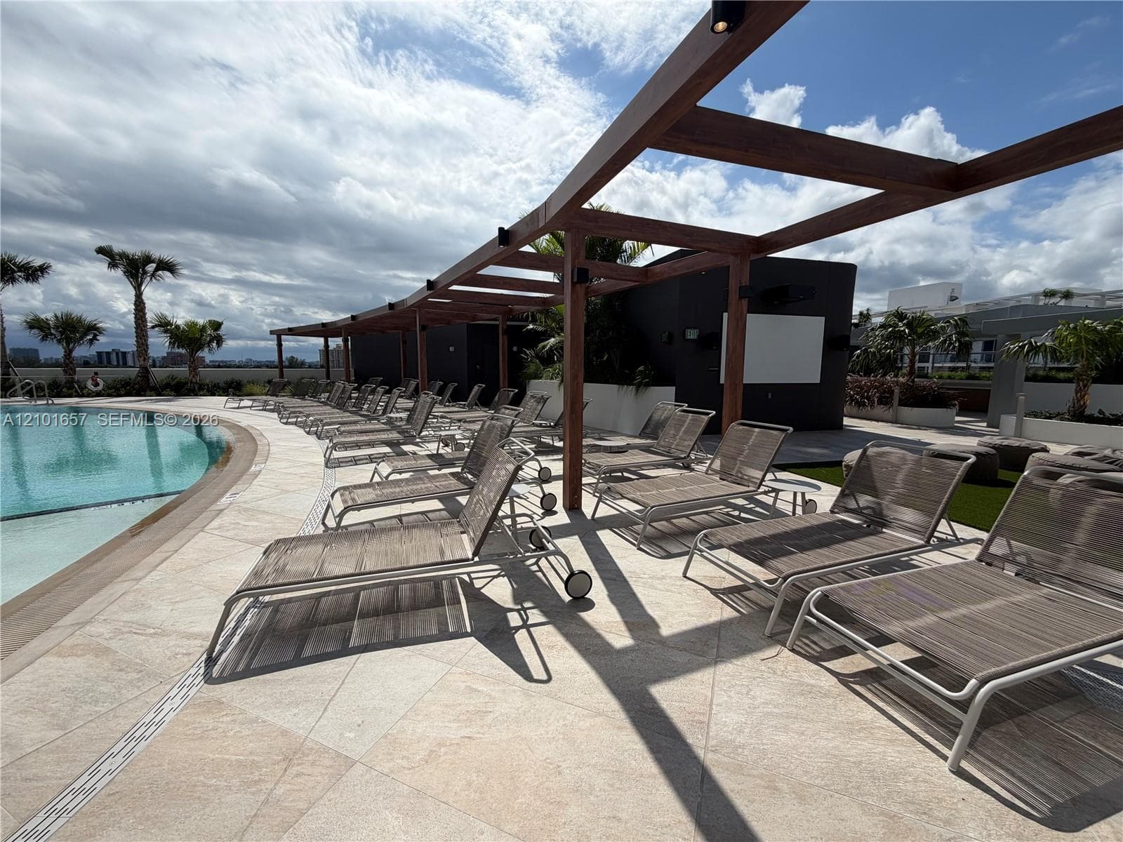 Curved swimming pool with stone-tiled decking and an overhead wooden pergola structure