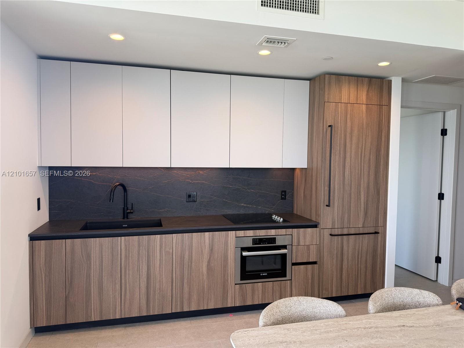 Modern kitchen featuring two-tone cabinetry with wood-finish lower cabinets, matte white upper cabinets, integrated refrigerator, stone backsplash, and recessed lighting