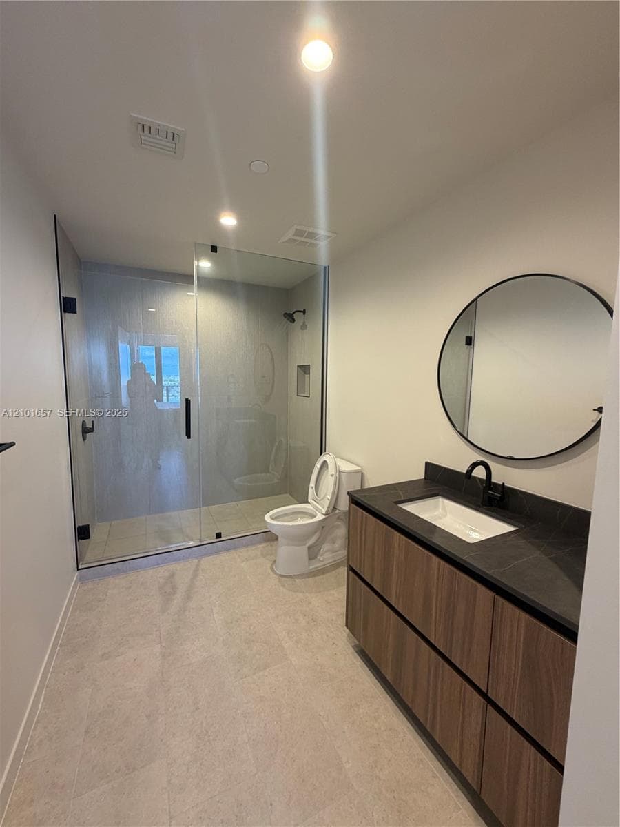 Floating vanity featuring wood-finish cabinetry, dark countertop, and integrated rectangular sink, paired with a frameless glass-enclosed shower, matte black fixtures, and a circular wall-mounted mirror