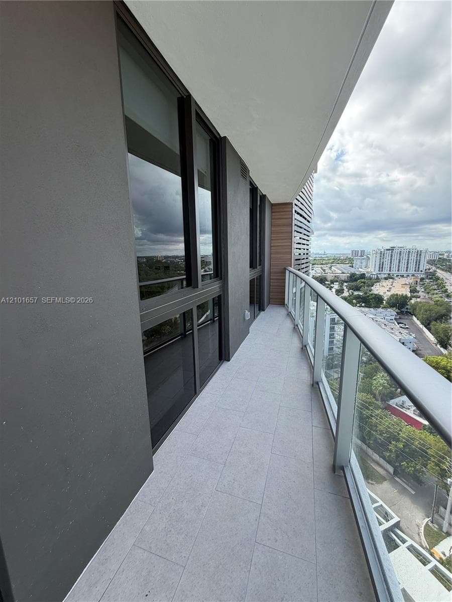 Private balcony featuring stone-finish tile flooring, glass panel railing with metal handrail, full-height windows, and wood-finish accent paneling