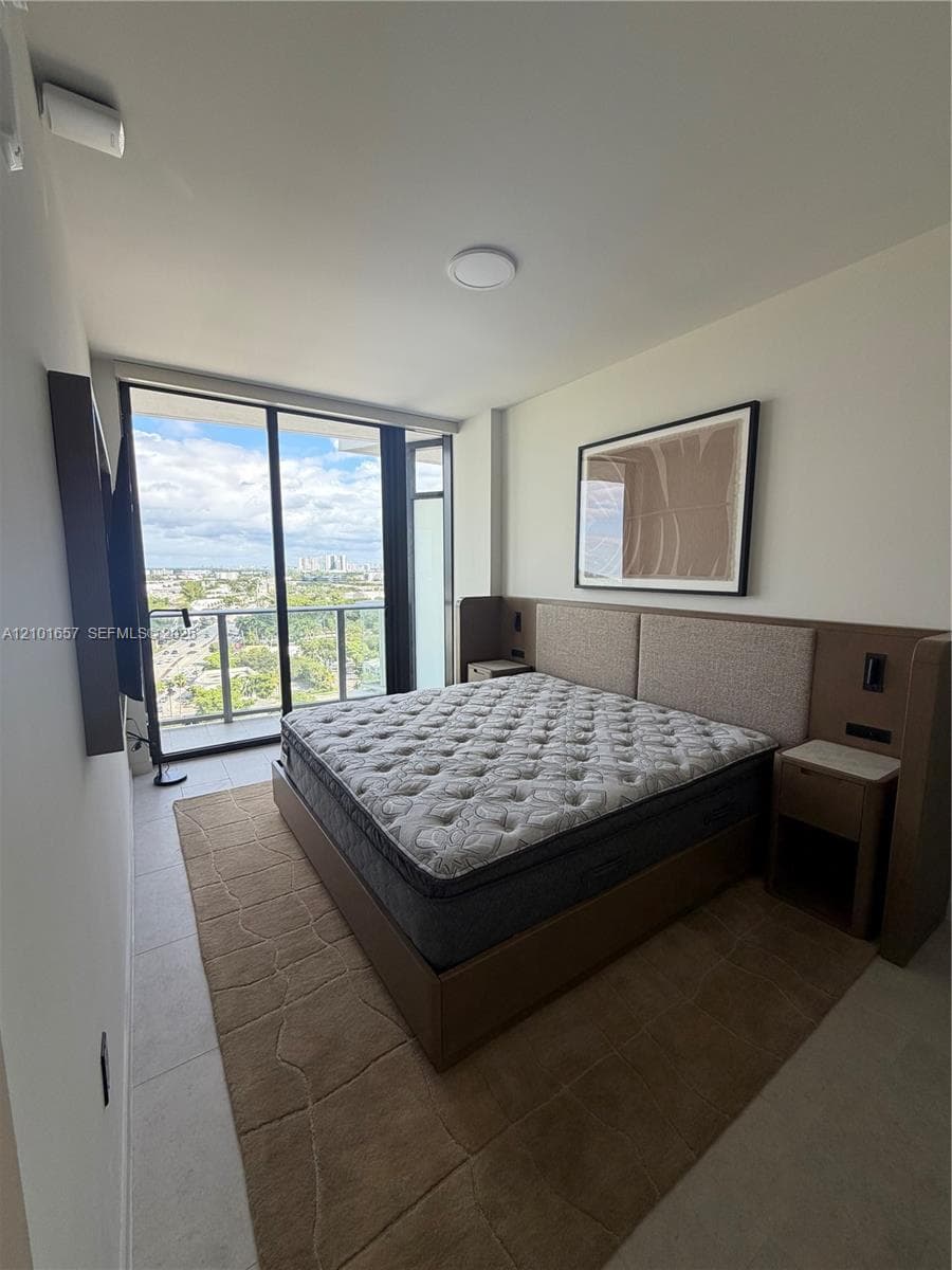 Floor-to-ceiling glass sliding doors opening to a balcony, tile flooring, and a built-in wood-finish headboard with integrated side tables
