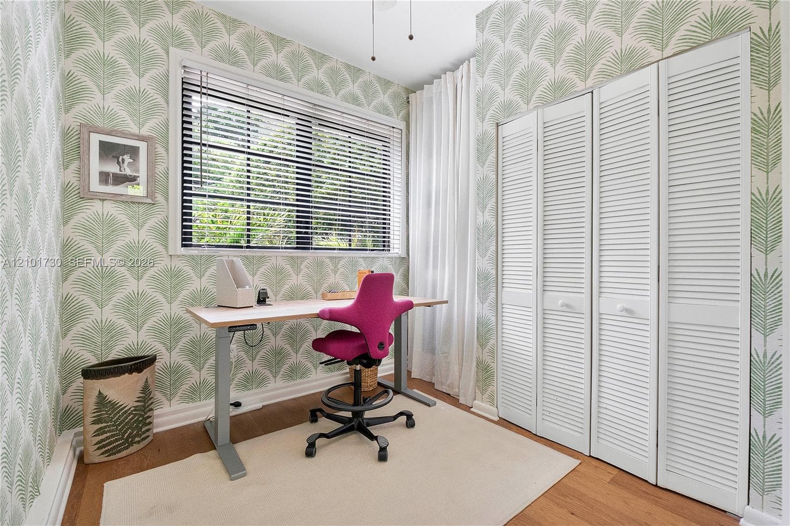 Room featuring palm leaf patterned wallpaper, wood-finish flooring, a multi-pane window with horizontal blinds, and a built-in closet with white louvered doors