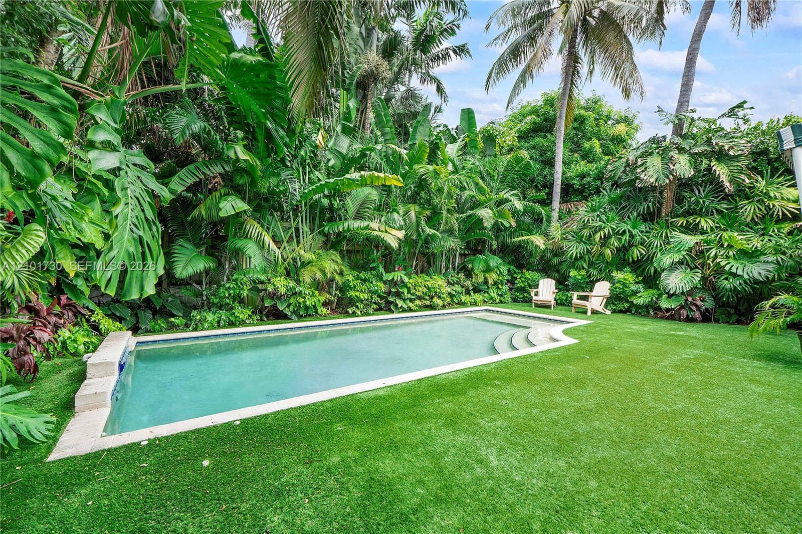 In-ground swimming pool featuring stone coping and built-in steps, surrounded by a lawn and dense tropical landscaping