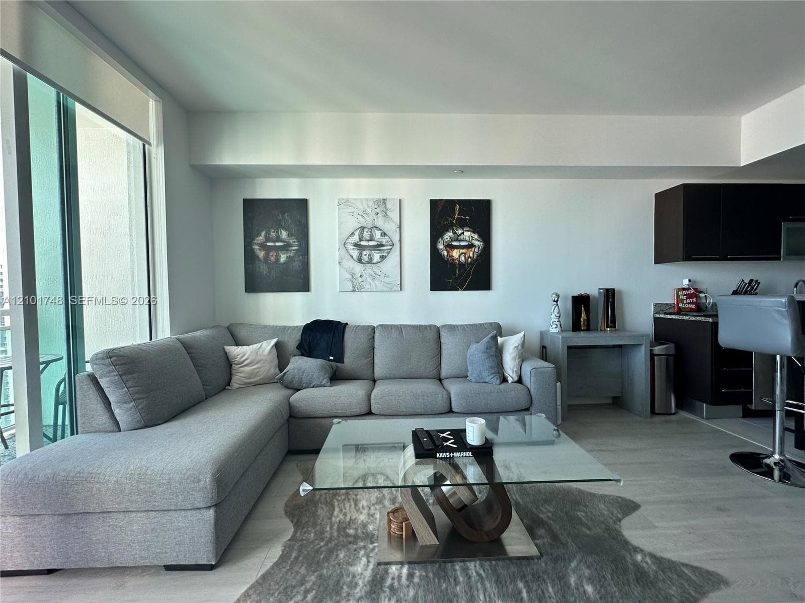 Open-concept living area with light-toned flooring, expansive glass doors, and a kitchen area featuring dark cabinetry and a stone-finish countertop