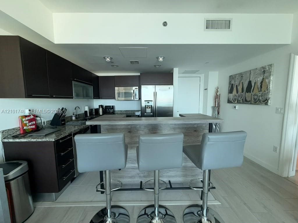 Kitchen featuring dark flat-panel cabinetry, stainless steel appliances, speckled stone countertops, and a wood-finish island with seating