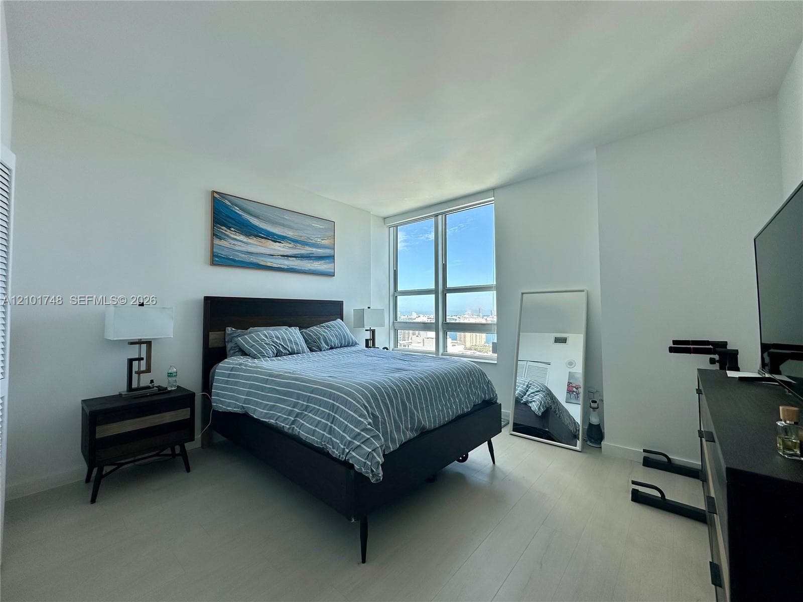 Expansive floor-to-ceiling window, light wood-finish flooring, white walls, and a built-in closet with louvered doors