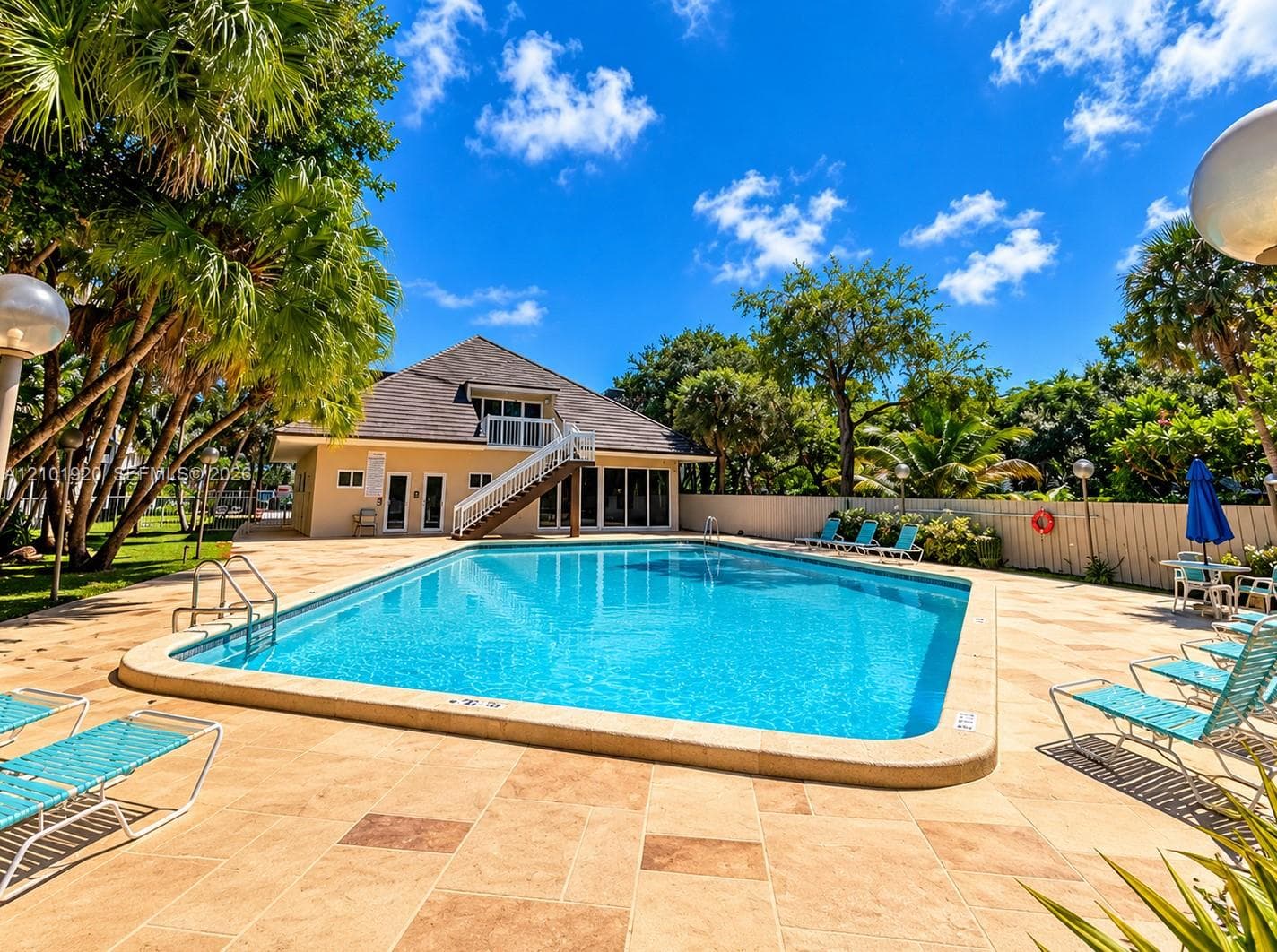 Outdoor swimming pool featuring a light-toned paved deck, metal pool ladder, and surrounding mature palm trees