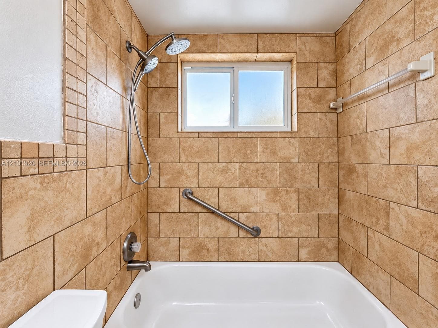 Full-height wall tile in a running bond pattern, white acrylic tub, adjustable dual-head shower fixture, built-in grab bar, and inset window