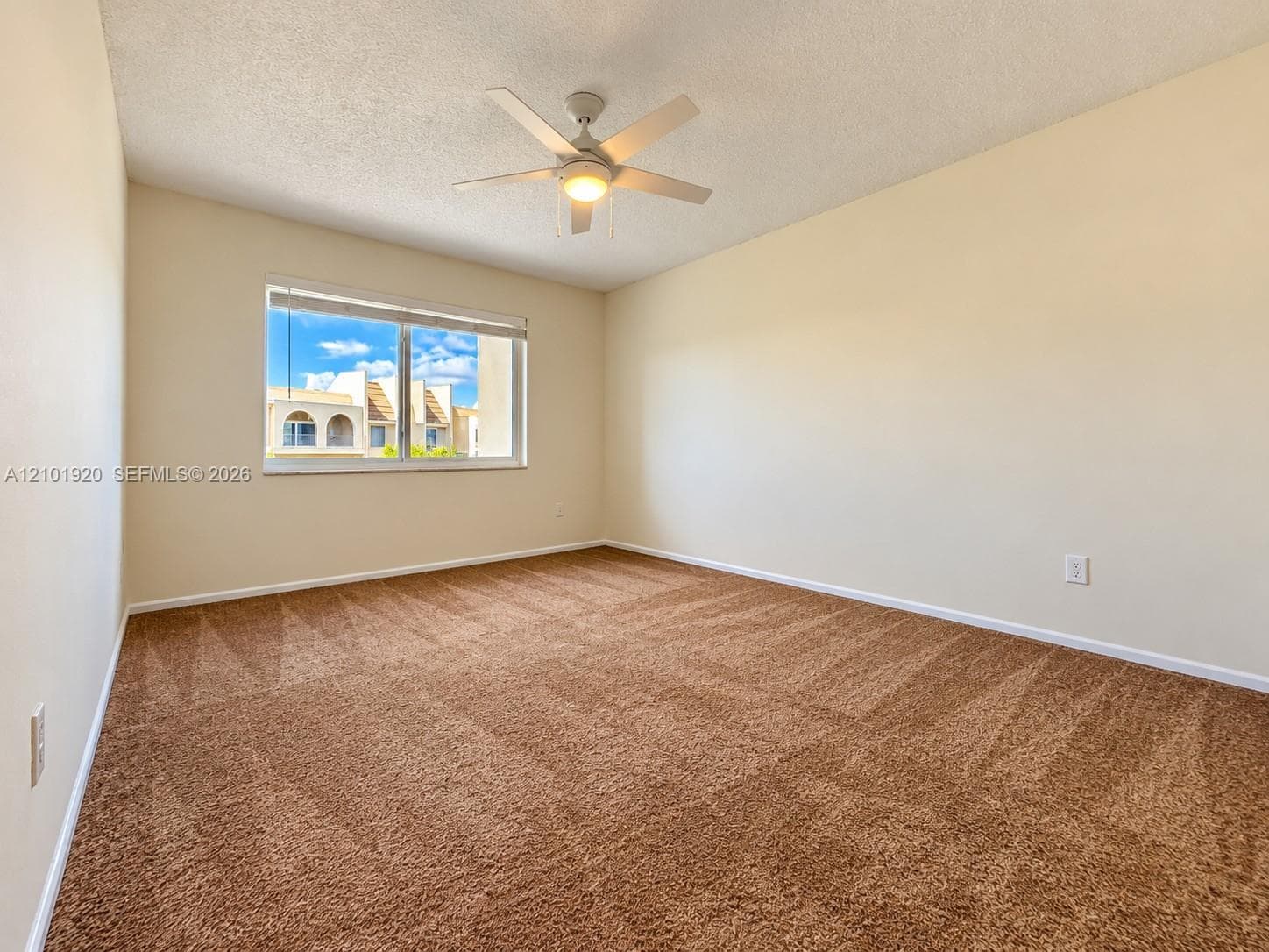 Neutral-toned interior space featuring textured carpeting, a white ceiling fan with integrated lighting, and a single-pane window with white horizontal blinds