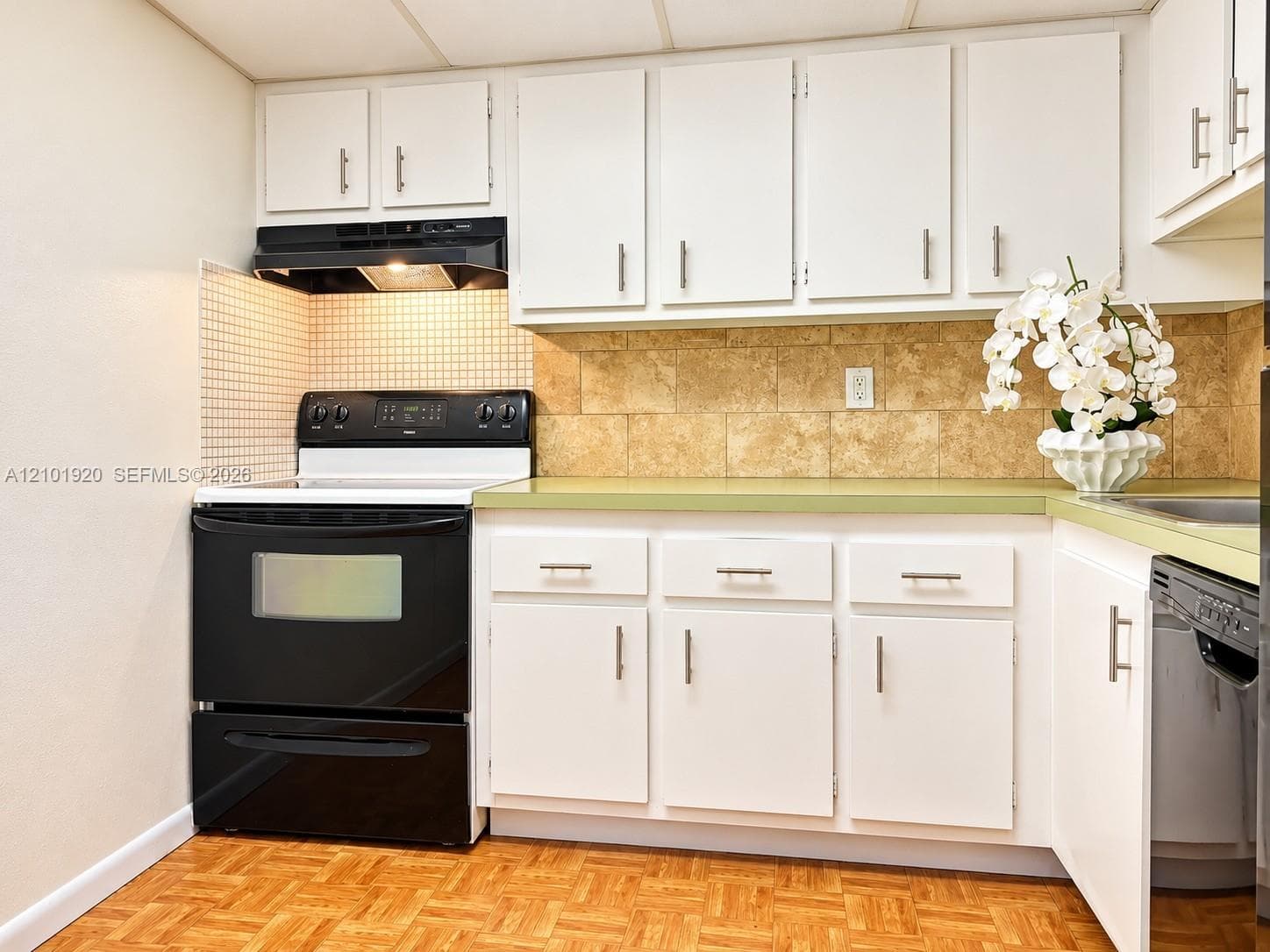 Kitchen featuring white shaker-style cabinetry with satin nickel hardware, light green countertops, stone-tile backsplash, and a black range with an integrated hood