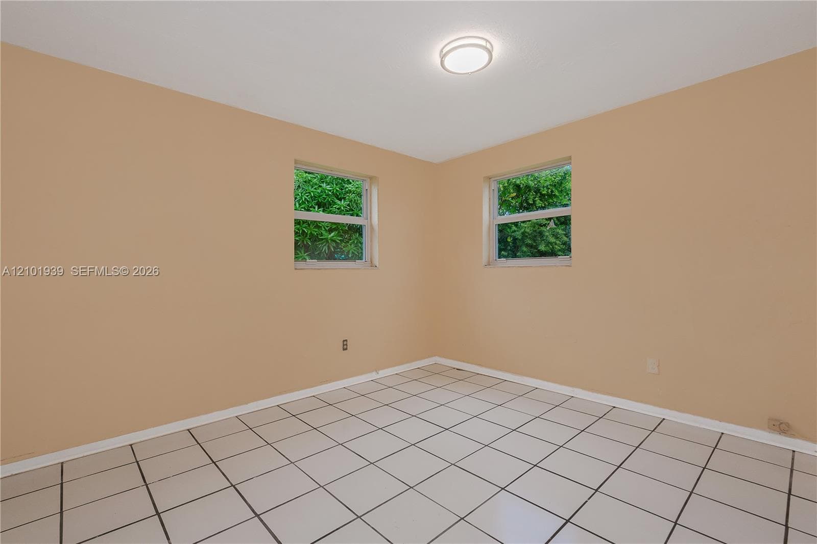 Neutral-toned interior space featuring light-colored ceramic tile flooring, two windows with exterior greenery, and a central flush-mount ceiling light fixture