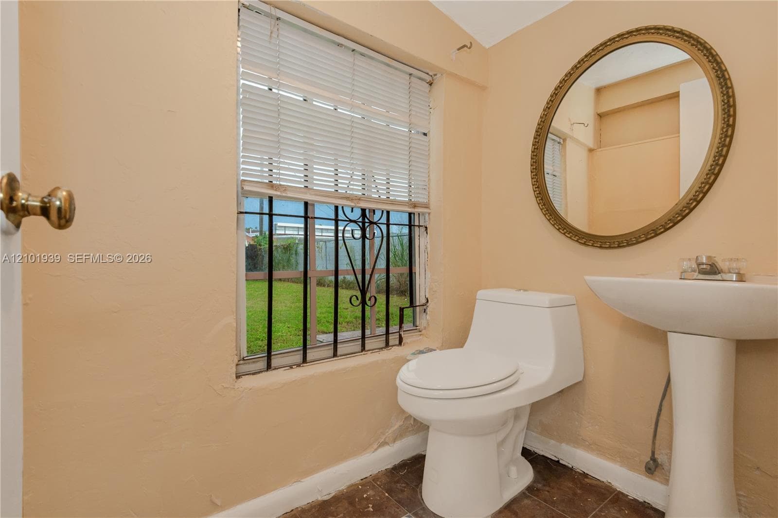 Window featuring iron security bars and horizontal blinds, pedestal sink, oval wall-mounted mirror, and low-profile toilet