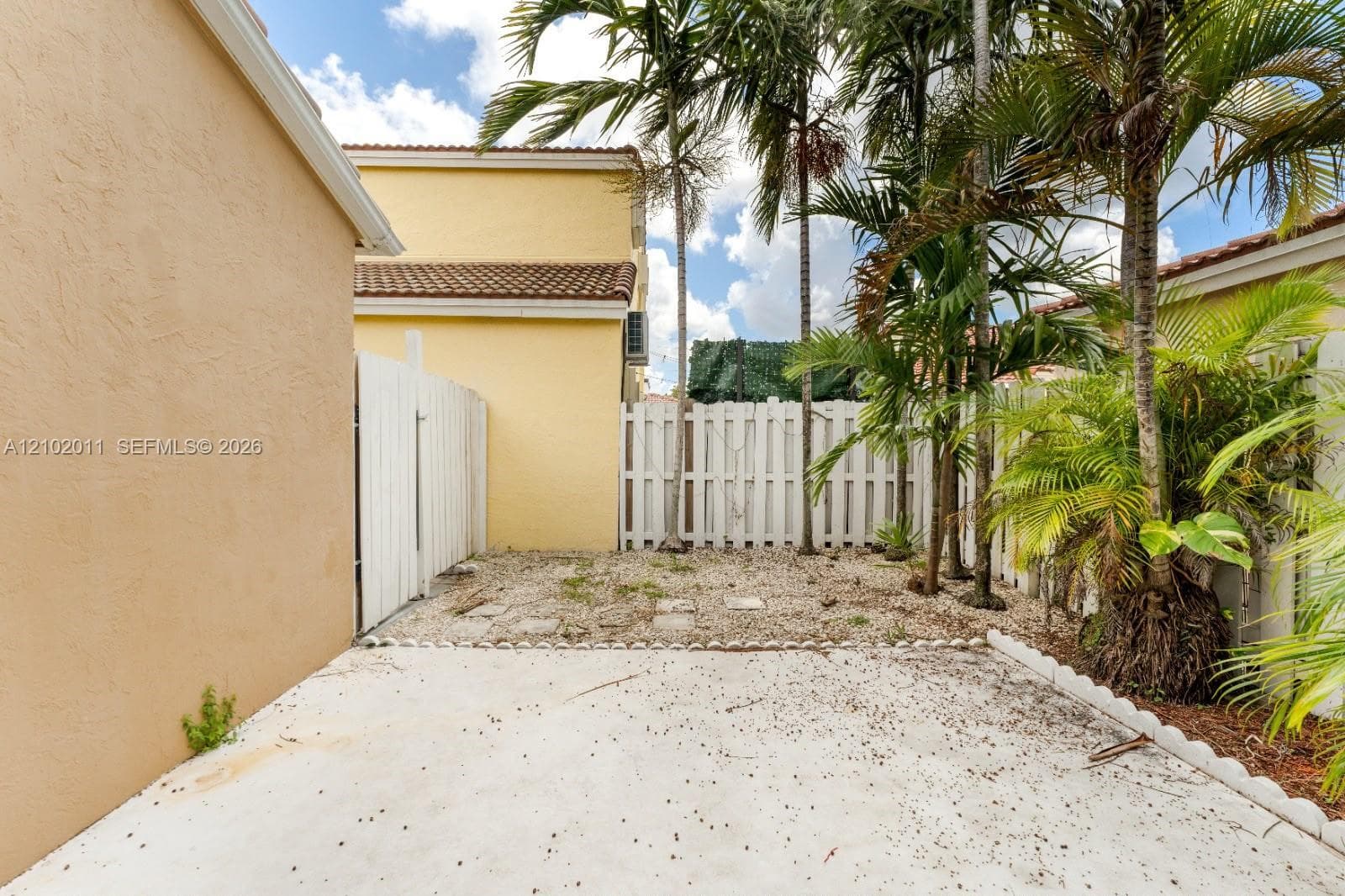 Paved concrete area featuring a white picket fence, mature palm trees, and exterior stucco walls