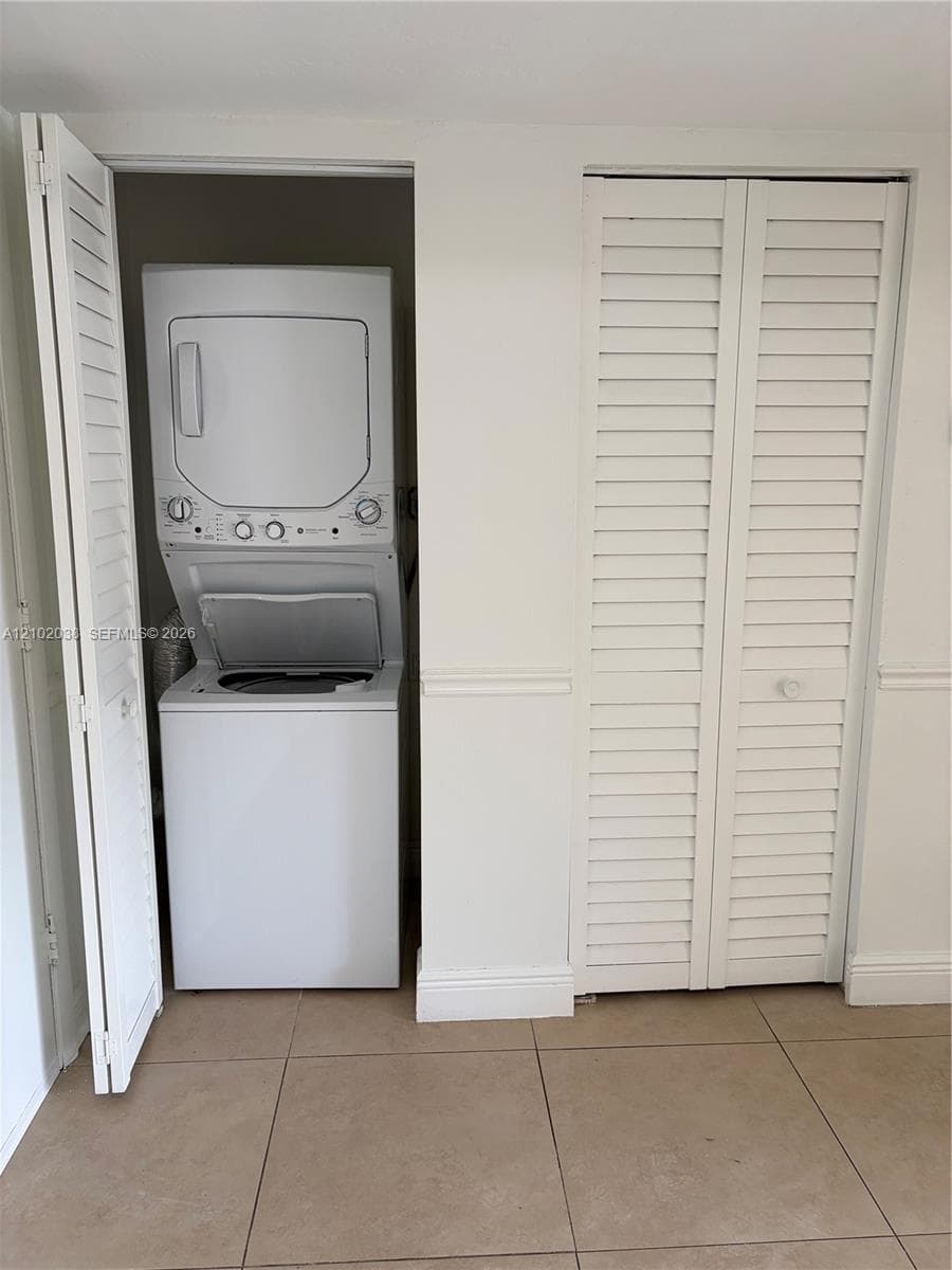 Laundry area featuring a stacked washer and dryer unit enclosed by white louvered doors, accented with white chair rail molding and tile flooring