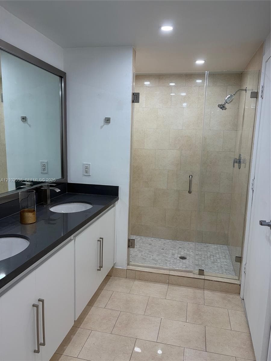 Dual-sink vanity with dark stone countertop, white cabinetry, large wall-mounted mirror, walk-in shower with glass enclosure, and stone tile flooring