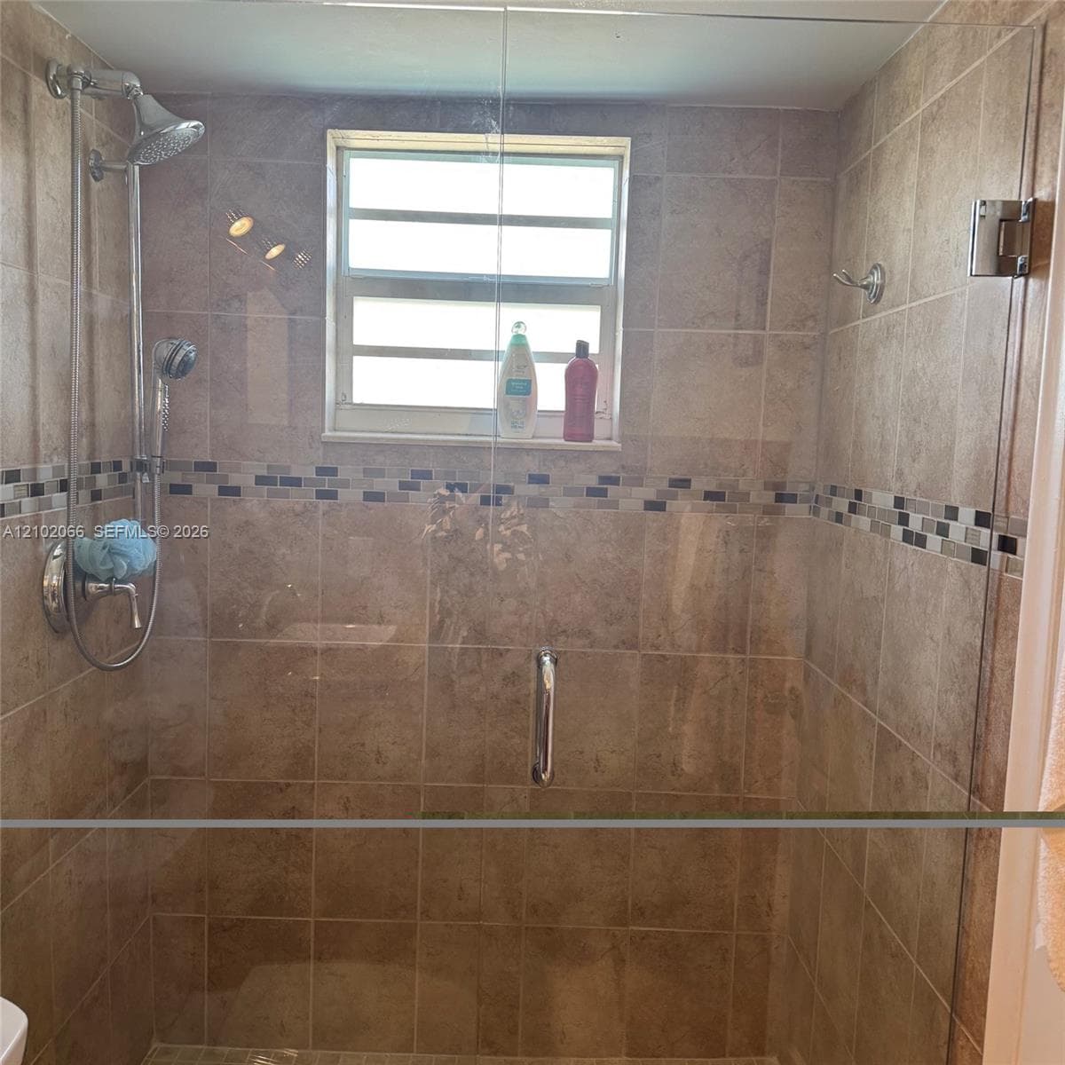 Walk-in shower featuring floor-to-ceiling neutral stone-tile surround, glass shower panel with polished chrome hardware, dual showerhead system, and an inset glass-tile decorative accent border