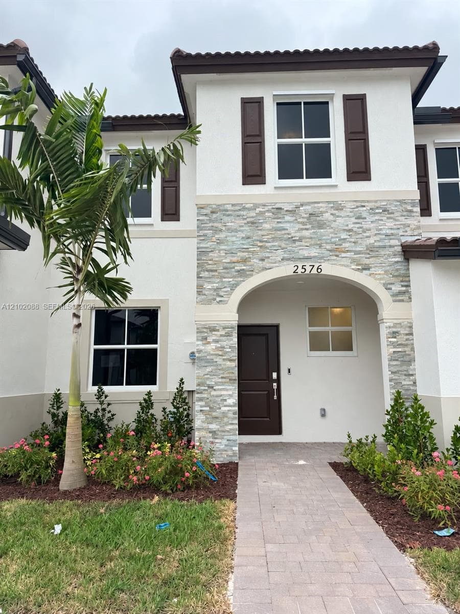 Stucco exterior featuring stacked stone accents, arched entryway, dark brown decorative shutters, and paver walkway