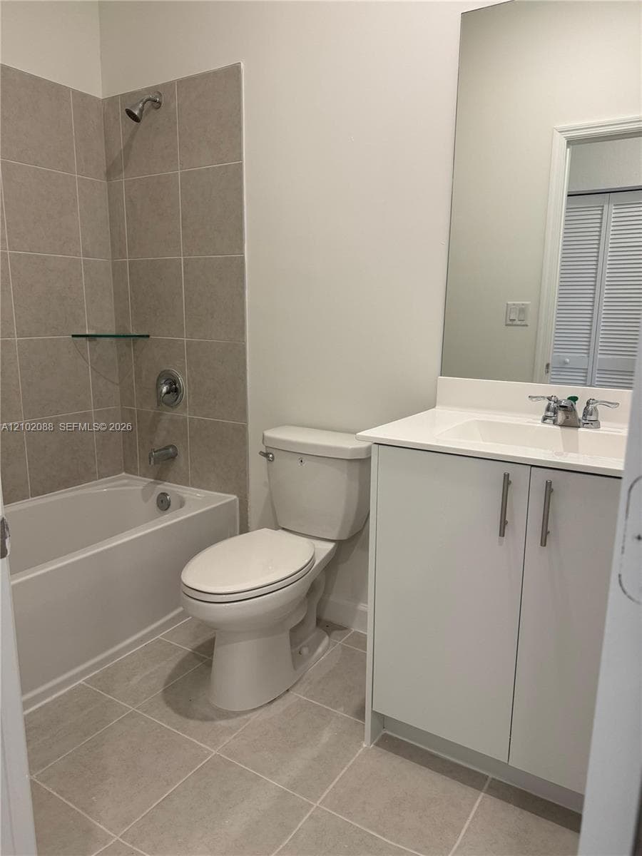 Bathroom featuring white cabinetry, integrated sink, large vanity mirror, porcelain tile flooring, and a tiled shower surround with a built-in glass shelf