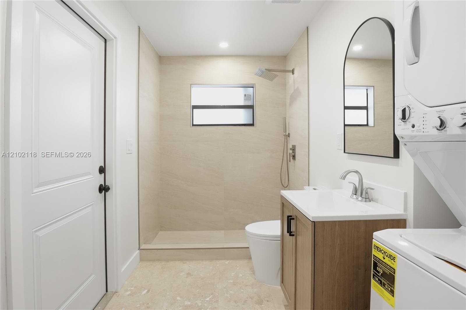 Walk-in shower with neutral stone-finish tile surround, wood-finish vanity with a white countertop, arch-top mirror, and light stone-finish flooring
