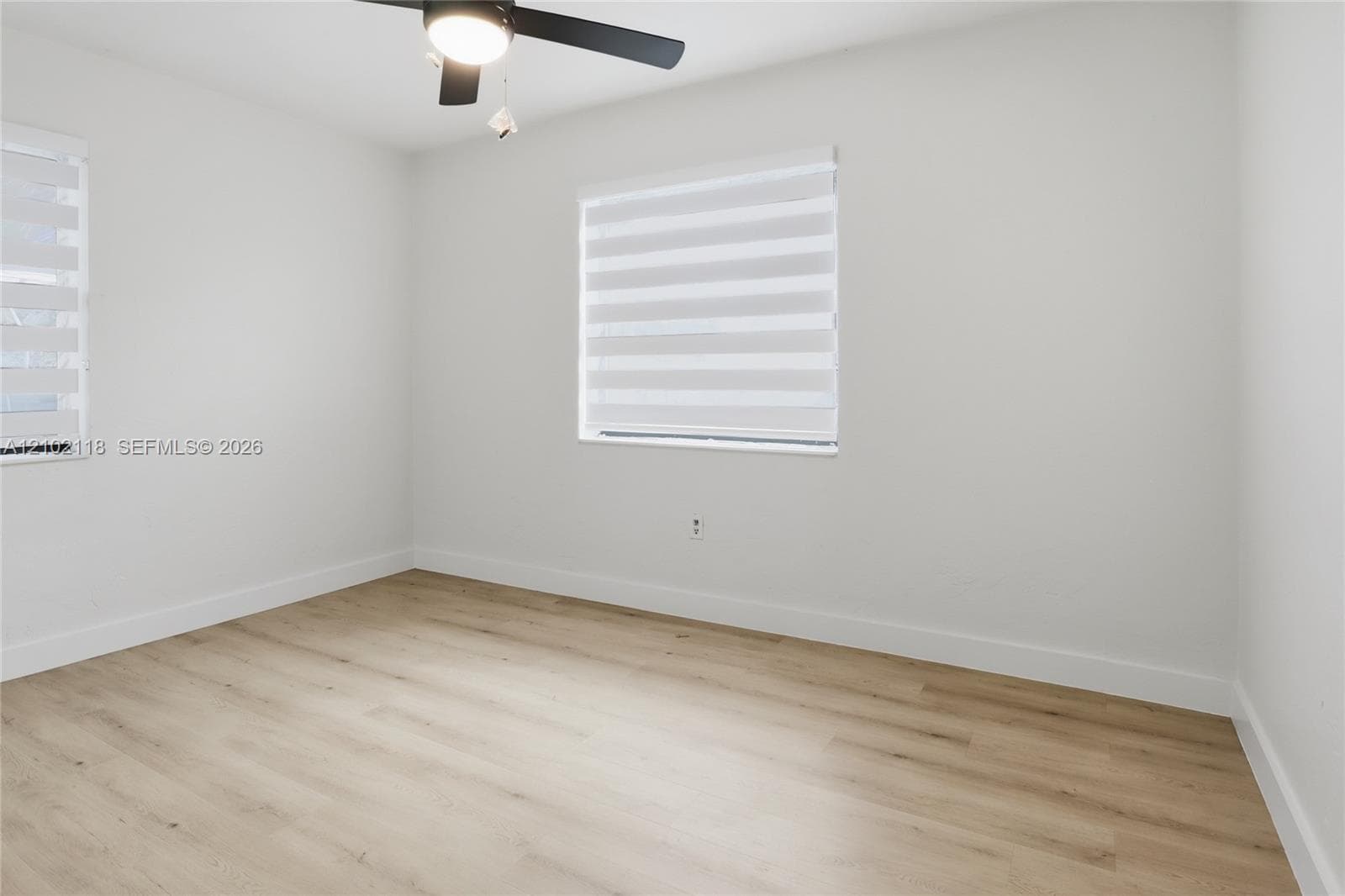 Interior room featuring wood-finish flooring, neutral wall tones, two windows with horizontal banded shades, and a ceiling-mounted light fixture with fan blades