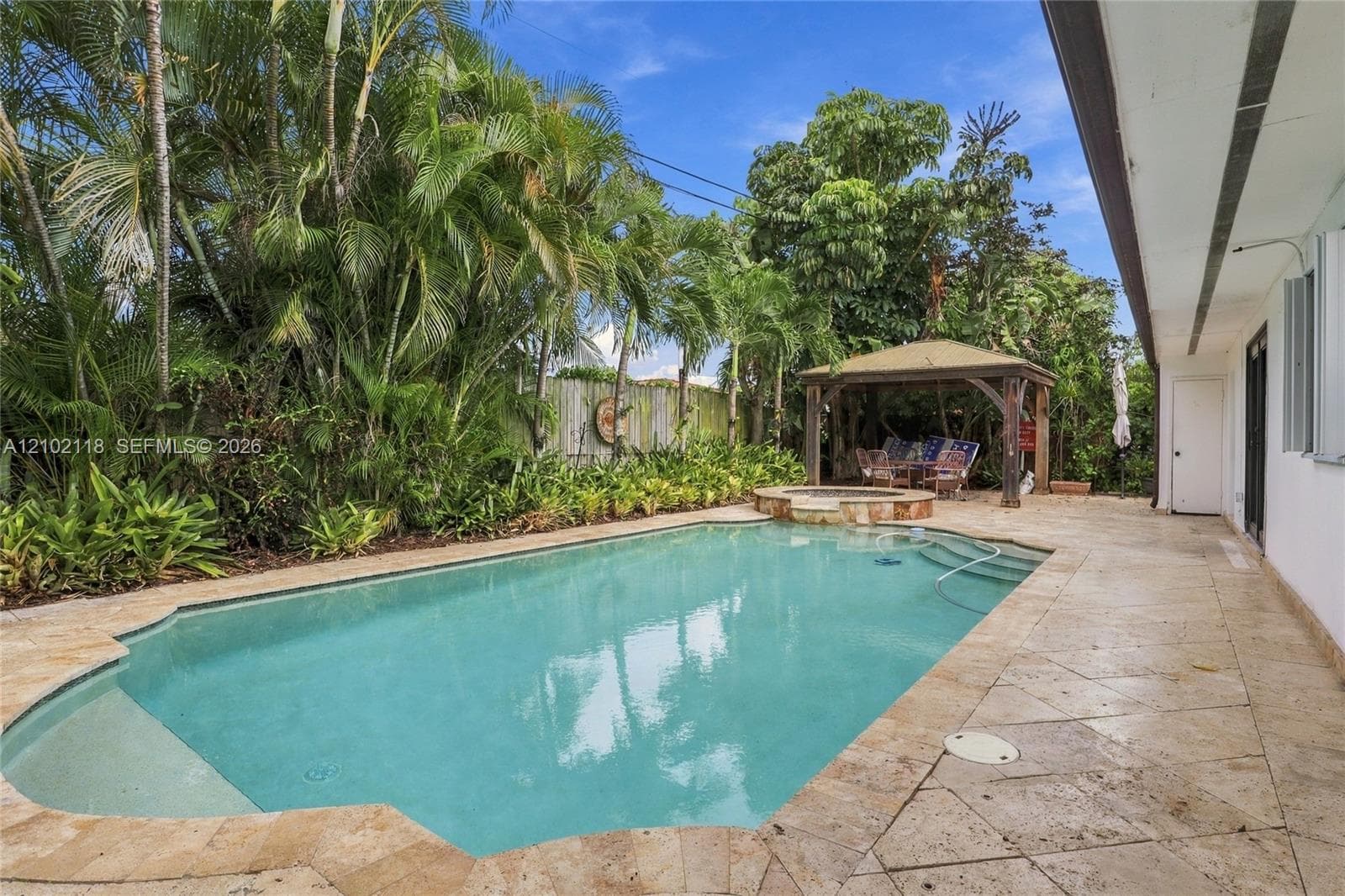 Outdoor area featuring a swimming pool with stone coping, a raised spa, natural stone paver decking, a wooden gazebo, and perimeter landscaping