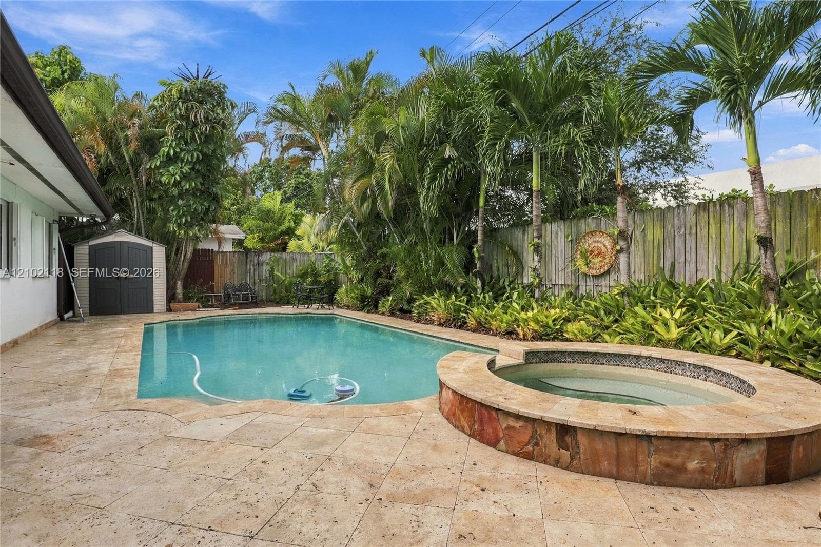 Backyard featuring a custom swimming pool, raised stone-finished spa, natural stone paver decking, privacy fencing, and mature palm trees