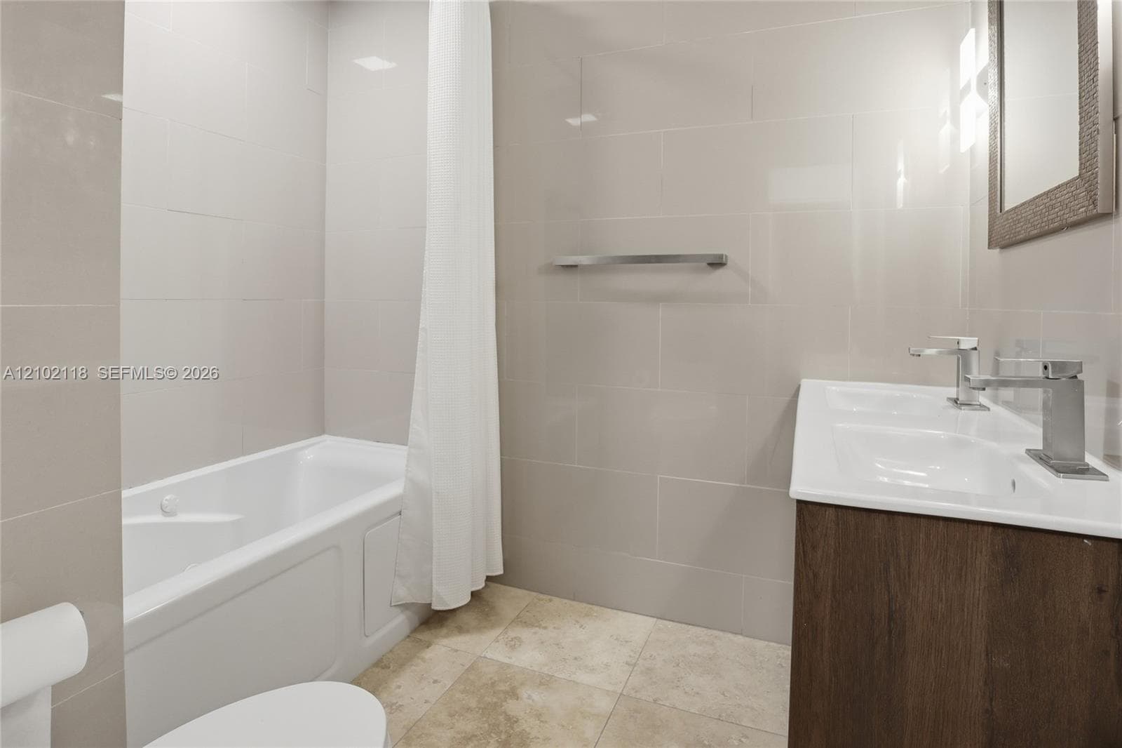 Bathroom featuring full-height neutral wall tiling, a soaking tub, a double-basin vanity with wood-finish cabinetry, and polished chrome faucets