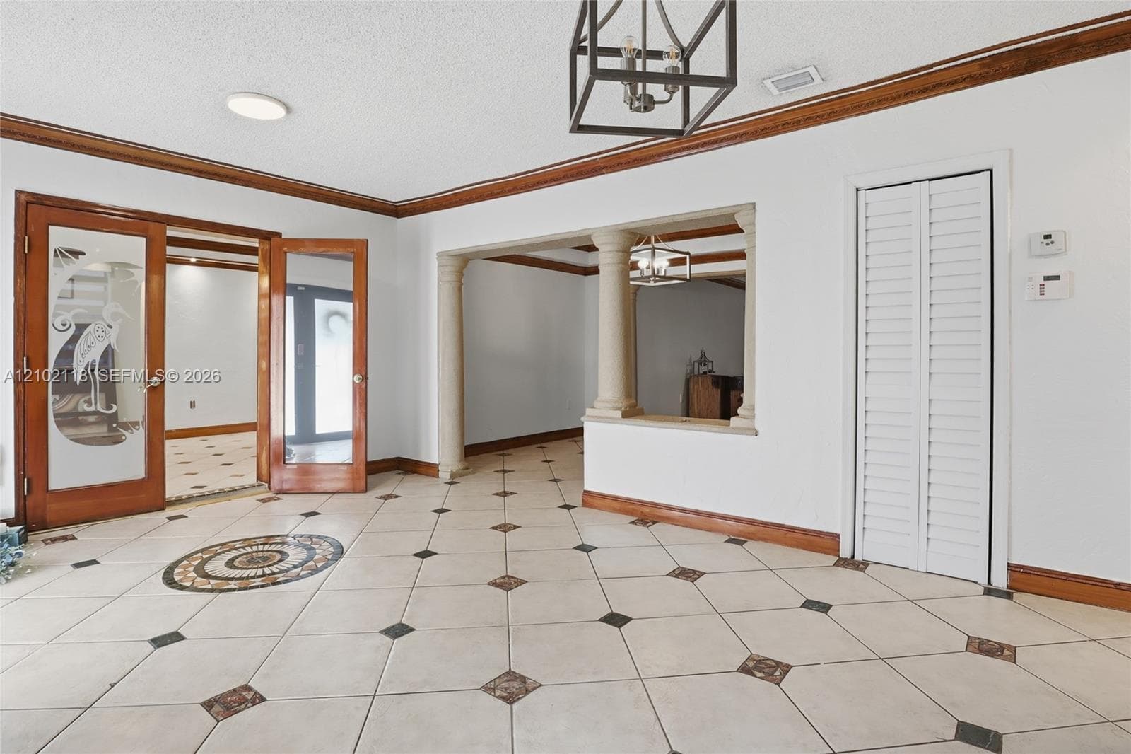 Ceramic tile flooring with decorative stone inlays, architectural columns, custom etched-glass double entry doors, wood-trimmed crown molding, and a built-in folding louvered closet door