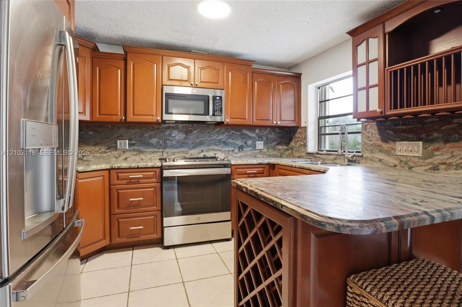 Kitchen featuring wood cabinetry, stone countertops with matching backsplash, stainless steel appliances, built-in wine rack, and tile flooring