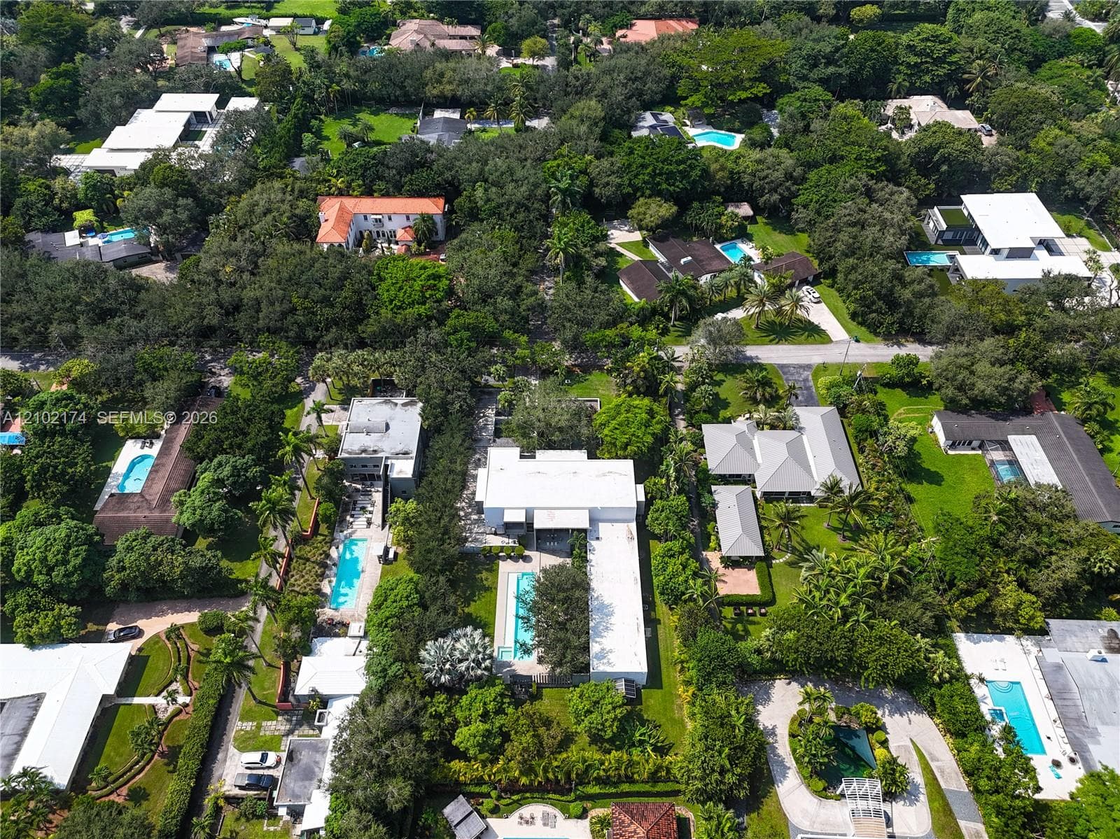 Aerial perspective featuring a flat-roofed estate with a rectangular swimming pool, expansive green lawn, mature trees, and paved driveway
