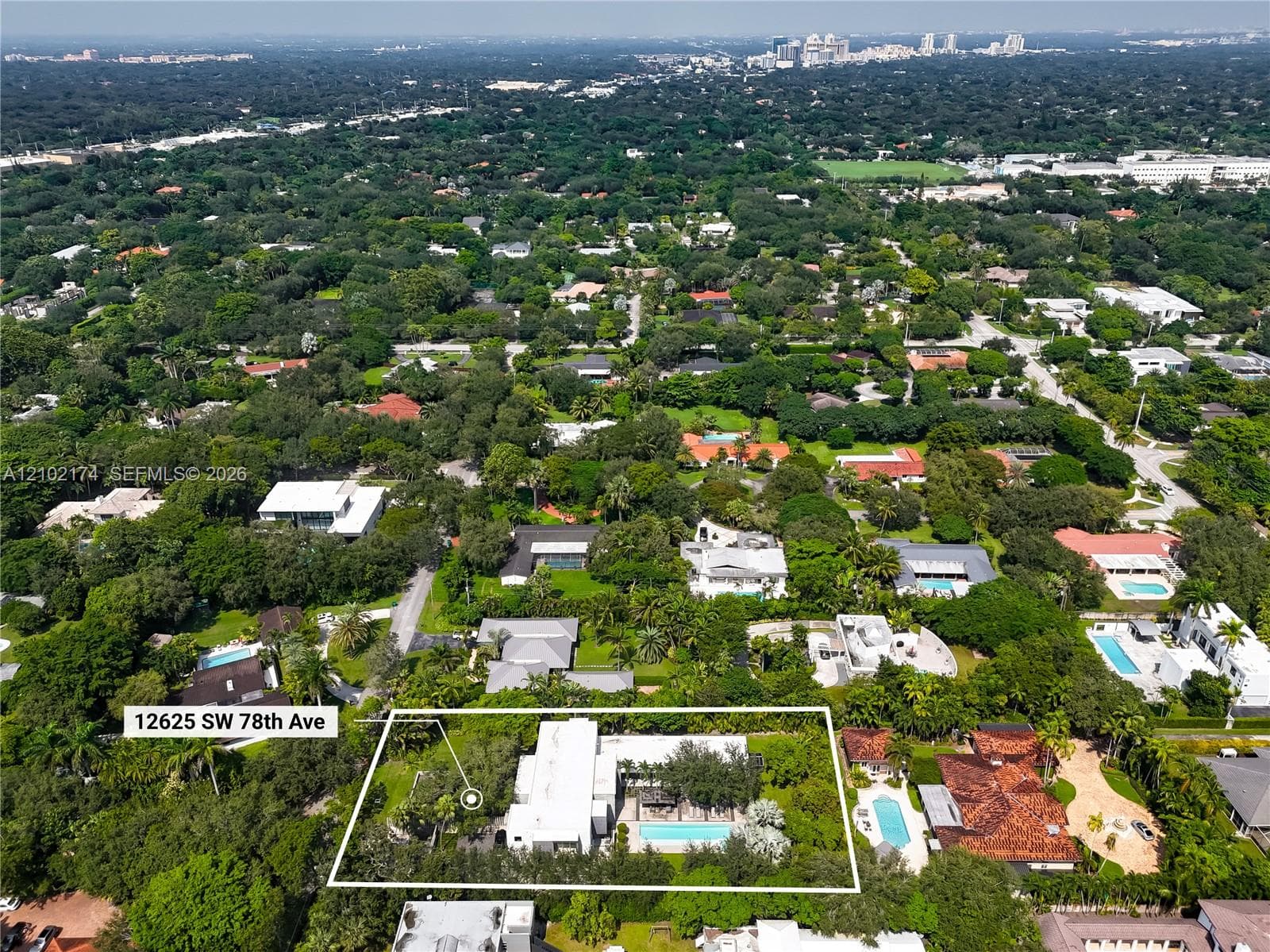 Aerial perspective of a property featuring a rectangular swimming pool, expansive manicured lawn, mature tree canopy, and multi-level roof structure