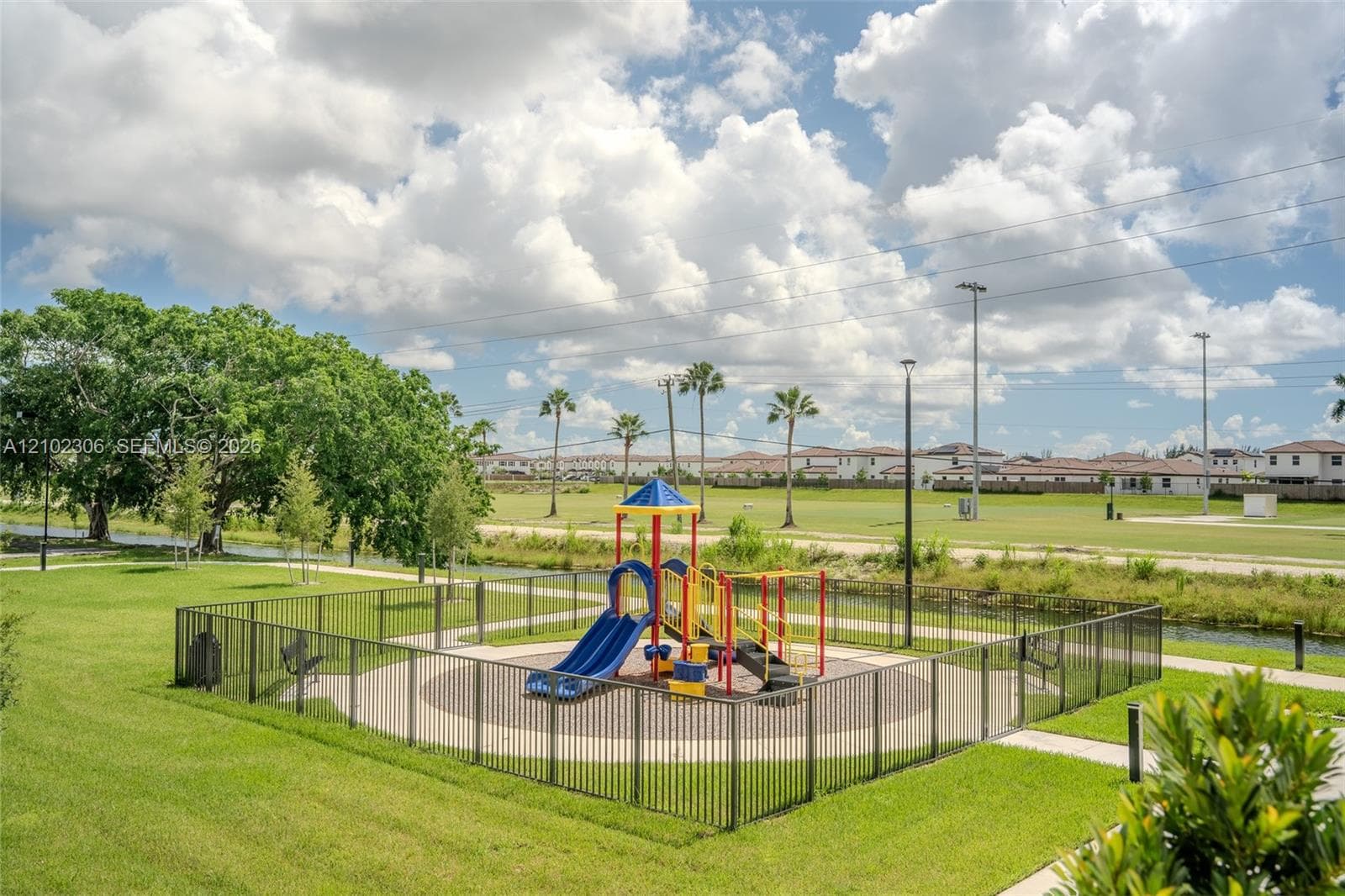 Outdoor recreation space featuring a fenced playground, manicured green space, paved pathways, and mature trees