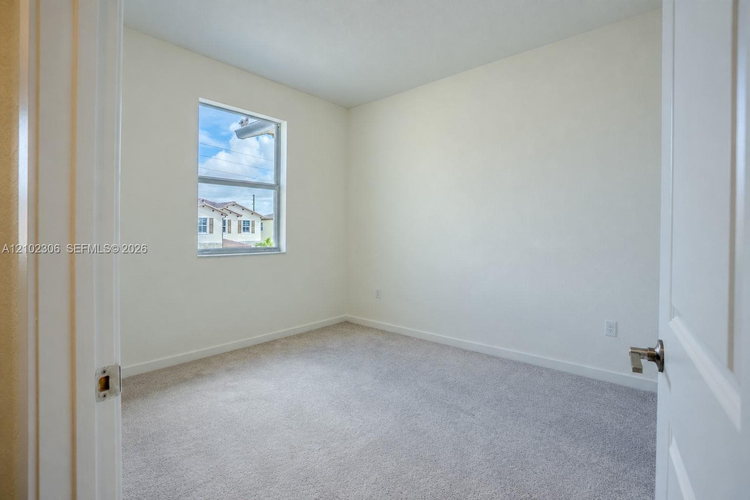 Neutral-toned interior space featuring light-colored wall-to-wall carpeting, a single-pane window, and a paneled interior door with a brushed nickel lever handle