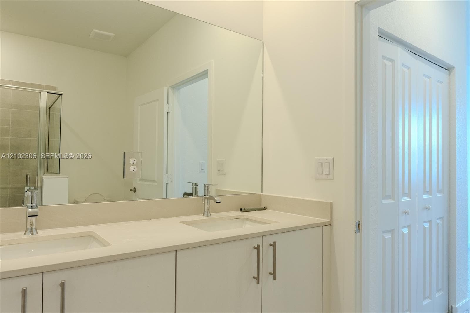 Dual-sink vanity with a solid-surface countertop, chrome faucets, floor-to-ceiling mirror, and white shaker-style cabinetry with bi-fold closet doors