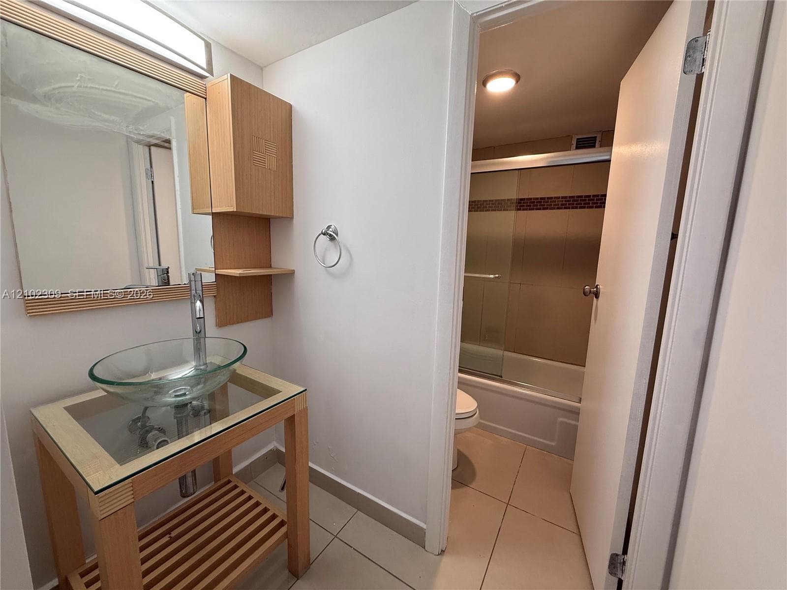 Bathroom featuring a light-wood vanity with a glass countertop, clear vessel sink, tall chrome faucet, and matching wall-mounted medicine cabinet, complemented by tile flooring and a tub-shower combination with a sliding glass door