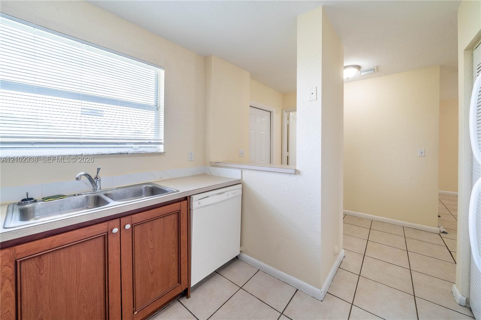 Double stainless steel sink with a window, white dishwasher, wood-finish cabinetry, and neutral tile flooring