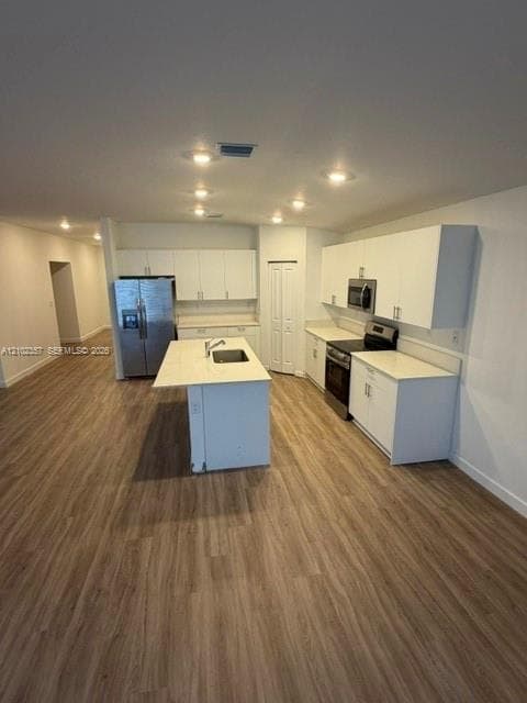 Kitchen featuring white cabinetry, solid-surface countertops, center island with an integrated stainless-steel sink, stainless-steel appliances, and wood-finish flooring