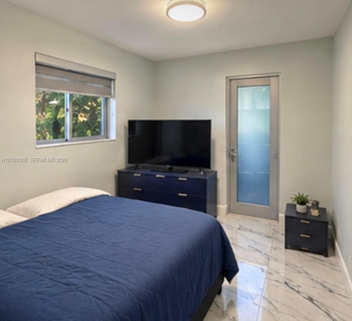 Room featuring large-format tile flooring with a marble-style finish, a single-pane window with a dual-shade treatment, a frosted glass-panel door, and a flush-mount ceiling light