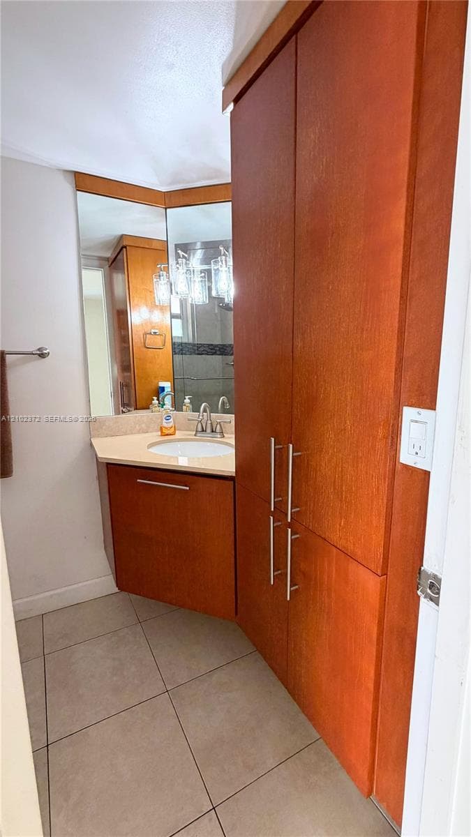 Custom wood-finish cabinetry, floor-to-ceiling storage unit with brushed metal hardware, solid-surface vanity countertop, drop-in sink with dual-handle faucet, and ceramic tile flooring